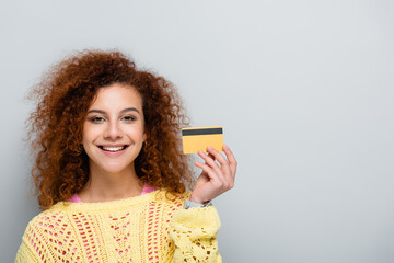 joyful woman in openwork sweater holding credit card isolated on grey - obrazy, fototapety, plakaty