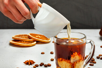 Human hand pours cream into a transparent cup with coffee, close-up.