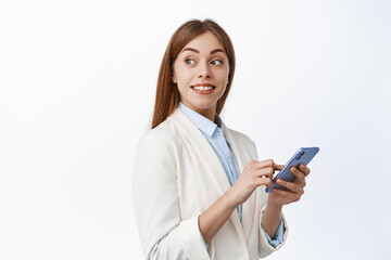 Smiling corporate girl in suit using mobile phone, turn head behind and look at promotional text, reading copy space on white background, holding smartphone