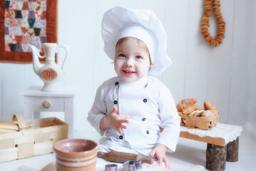 a little girl, blonde, in a white chef's hat, smeared in flour, with bagels around his neck, kneads dough in a brown wooden kitchen, prepares bread and pasta and tries everything