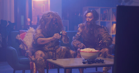 Prehistoric couple of cavemen friends entering dark smoked house living room watching football and eating popcorn like modern people. Entertainment.