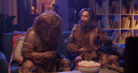 Prehistoric couple of wild people in fur clothes drinking beer throwing eating popcorn having fun in modern living room house. Entertainment. Movie concept.