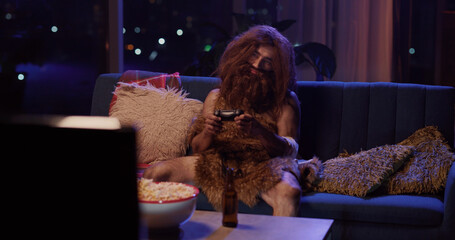 Bearded ancient savage man in furry animal leather clothes falling asleep after playing videogames on television in modern house. Humor concept.