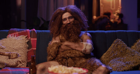 Portrait of funny ancient prehistoric primitive human in animal fur covered sitting on sofa...