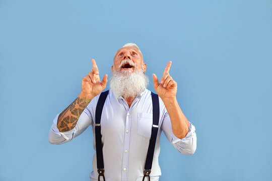 Fashionable Senior Man With Grey Hair And Long Beard Wearing Suspenders Looking Up Amazed And Surprised.