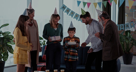 Cheerful diverse family celebrating birthday party. Happy parents and kids, their grandparents dancing in living room blowing candles on cake enjoying.