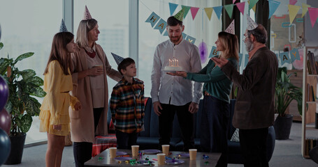 Cheerful diverse family celebrating birthday party. Happy parents and kids, their grandparents dancing in living room blowing candles on cake enjoying.