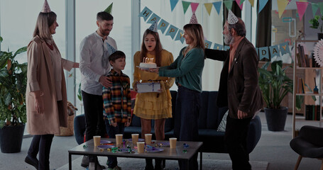 Caucasian family members congratulating teenager beautiful happy girl with birthday, blowing candles on cake and sharing presents.