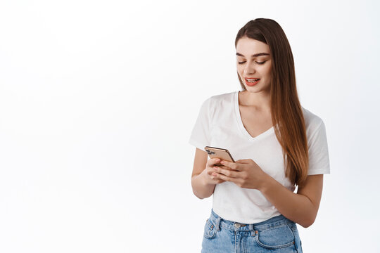 Technology and people concept. Modern girl using smartphone, texting, reading message on phone screen with casual relaxed face, standing in t-shirt against white background