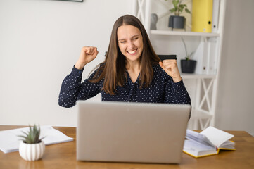 Excited woman sits in front of the laptop and rejoices, happy with good news female screams yes on the workplace, raised fists up, celebrates victory