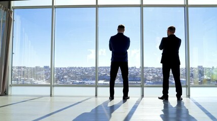 Meeting of entrepreneurs in office by the window. Rear view of two businessmen admiring the city landscape through the office window and talking about new project.