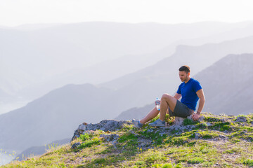 Naklejka premium Handsome athlete sitting at a rocky peak while looking at the breathtaking mountain line and a beautiful lake while wearing a blue shirt and grey shorts.