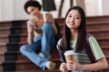 Cheerful asian student with coffee to go and notebook looking at camera