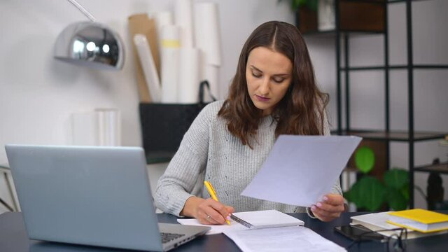 E-learning concept. Concentrated female student takes notes, writes down in notepad watching online webinars, video classes, studying from home, young woman doing paperwork
