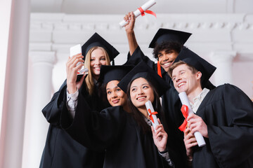 Interracial graduates with diplomas taking selfie in university
