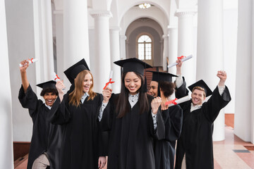 Asian graduate with diploma showing yes gesture near excited friends