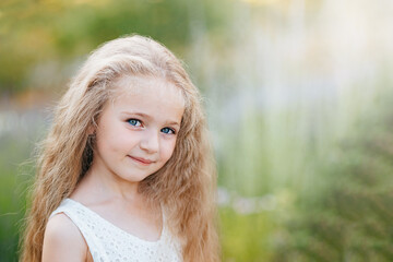 Close up portrait of pretty, blue eyed, fair skinned girl with happy and peaceful expression and long blonde curls, outdoors, backlight on bright sunny day. Cute child with dimpled cheeks