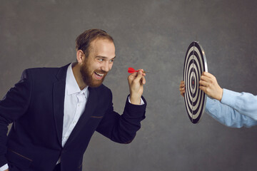 Ambitious businessman aiming red dart at target dartboard determined to throw it and hit bulls eye,...