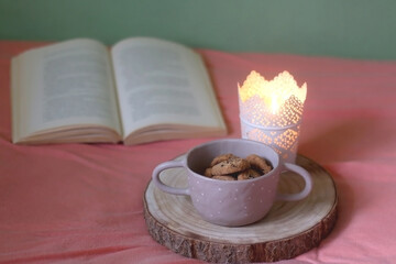 Bowl of chocolate chip cookies, lit candle and open book on a bed. Selective focus.