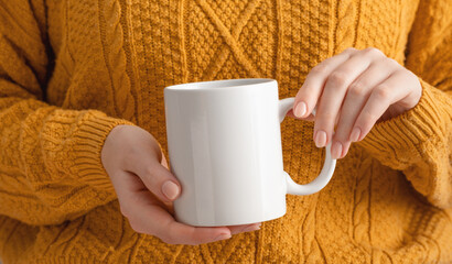 Female hands hold mock up white empty mug, cup for your design and logo close-up. Woman in yellow...