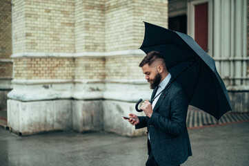 Businessman using a smartphone and holding a black umbrella outside