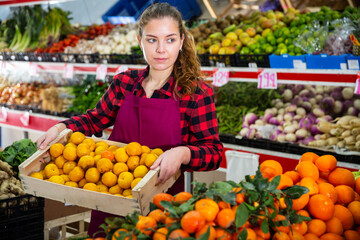 Portrait of a young saleswoman standing with a box of tangerines near the counter in a store