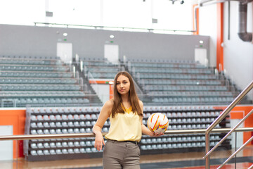 portrait of a young pretty woman with blue eyes and long hairs dressed in yellow blouse and trouses holding football ball in hands in white, orange and gray sport gym