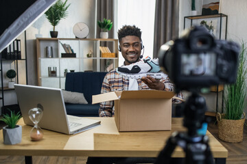 Happy young blogger doing live stream while unpacking new trendy sneakers. Afro american man using headset, video camera and laptop for his hobby.