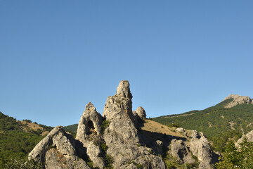 sharp mountain peaks with a cave on the background of a clear blue sky and a large number of green trees
