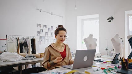 Young female fashion designer video chatting with client, showing color swatches to laptop, working at workshop - Powered by Adobe