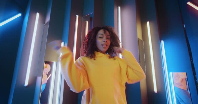 Active urban african american black woman in yellow hoodie dancing by neon lights background in disco club. Cool talented mixed race female dancer feeling free moves rhythmically to hip-hop music beat