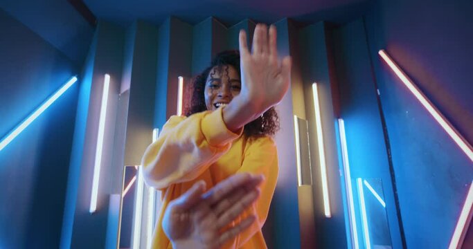 Cheerful african american woman in yellow hoodie dancing modern dance with hands by neon lights in disco club at night. Cool mixed race female feeling free moves rhythmically to hip-hop music beat