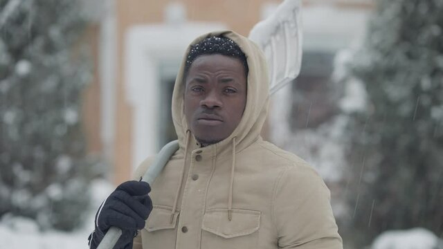 Portrait of perspiring African American man wiping forehead after cleaning snow at backyard and looking at camera. Tired handsome young guy posing at home in garden with shovel on shoulder