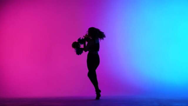 Silhouette of slender cheerleader, dancing energetically with pompoms in hands against background of pink blue lights in studio. Cheering dance in sports competitions. Slow motion.