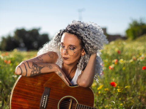 Cute Spanish woman with white curly hair and cool tattoos sitting in a meadow with a guitar