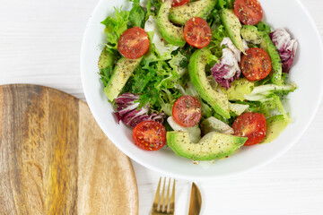 Fresh green salad with avocado, tomatoes, goat cheese in big white bowl on wooden tray. Vegan salad with green mix leaves  and vegetables. Top view on gray table. Healthy vegetarian food. Close up