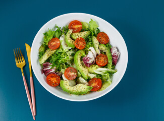 Fresh green salad with avocado, tomatoes, goat cheese in big white bowl on wooden tray. Vegan salad with green mix leaves and vegetables. Top view on blue table. Healthy vegetarian food. Close up	