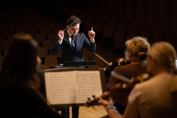 Conductor of symphony orchestra with performers in background in concert hall - obrazy, fototapety, plakaty