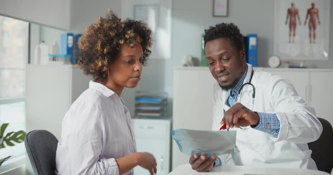 African male therapist in coat and showing x-ray and explaining problem to patient in modern office