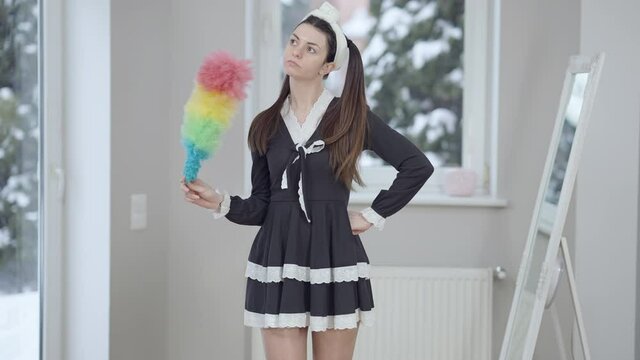 Slim beautiful Caucasian woman standing with colorful dust brush indoors thinking. Portrait of thoughtful charming millennial in maid uniform looking at camera and smiling