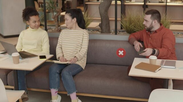 High angle of young Caucasian man using smartphone, then starting conversation with two beautiful Asian and Mixed-Race women sitting on couch on distance from him in restaurant
