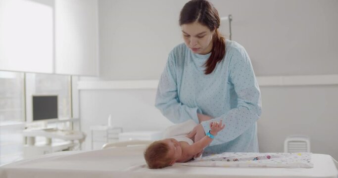 Young mother swaddling baby with white blanket on table in hospital
