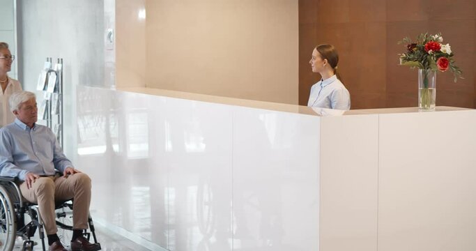 Professional receptionist talking to senior woman and handicapped man at desk in clinic