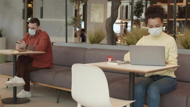 Medium-long of young Mixed-Race woman using portable computer, blurred man using smartphone sitting on couch in safe distance, people wearing masks in coworking place