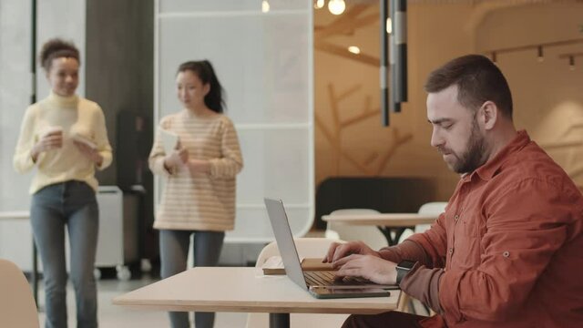 Side view of young Caucasian male restaurant manager sitting at table, typing on laptop computer, showing come here gesture with hand to young Asian and Mixed-Race women joining him for interview