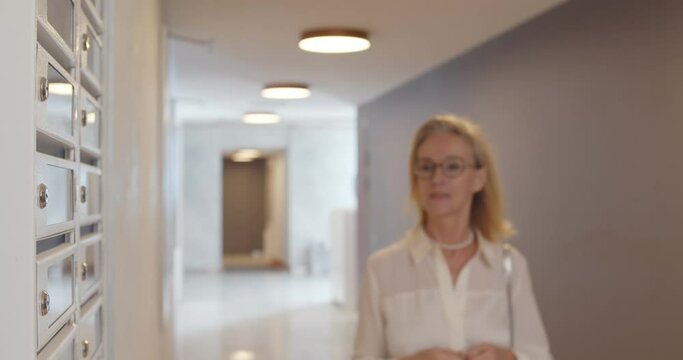 Senior female in eyewear opening postbox standing in hallway and holding envelope with invitation.