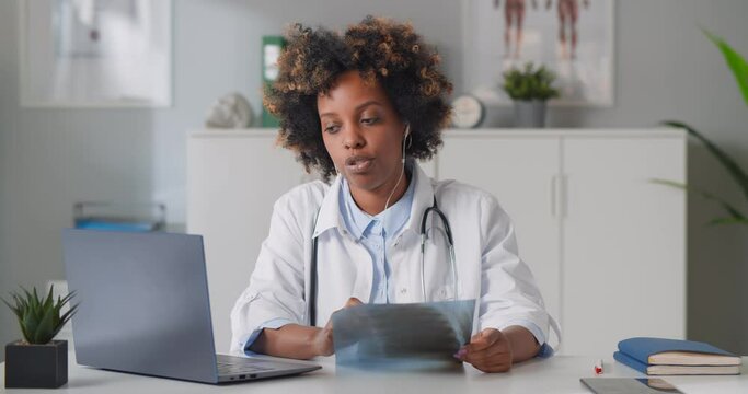 Female african doctor holding chest x-ray film consulting patient online on laptop