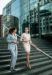 Two smiling business women talking while walking down stairs near modern office building.