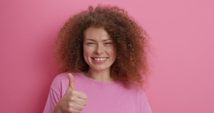 Pleasant looking cheerful woman looks at promo offer shows thumb up in aproval says very good satisfied with your choice smiles happily wears casual t shirt isolated over pink studio background