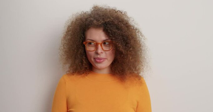 Positive good looking Caucasian woman bites ips winks eyes lookswith cheerful tricky expression wears transparent glasses orange jumper isolated over white background. People emotions concept
